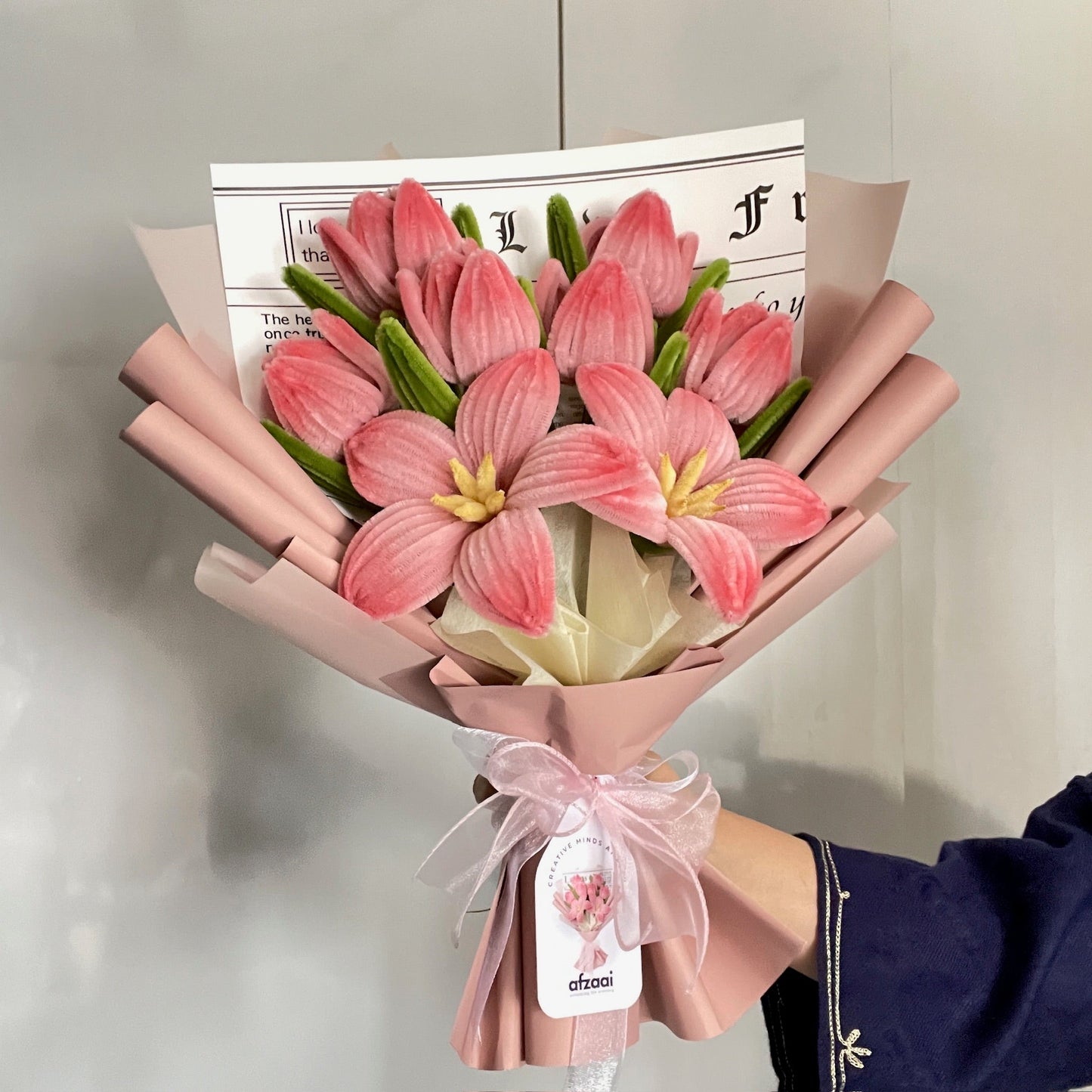A bouquet of six tulips and two lilies made using peach pink color pipe cleaners with a afzaai tag on a neutral background